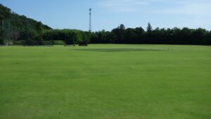 The Sano International Cricket Ground in Tochigi Prefecture, Japan, an open grass ground with net practice and a mobile sightscreen beside wooded hills